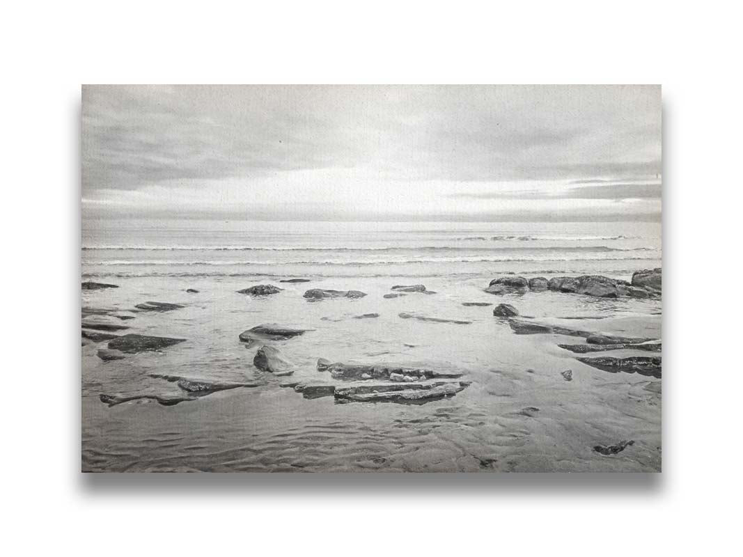 A grayscale photograph of the rocky shore of the North Sea at low tide. The sun peeks over the horizon through the partly cloudy sky. Printed on canvas.