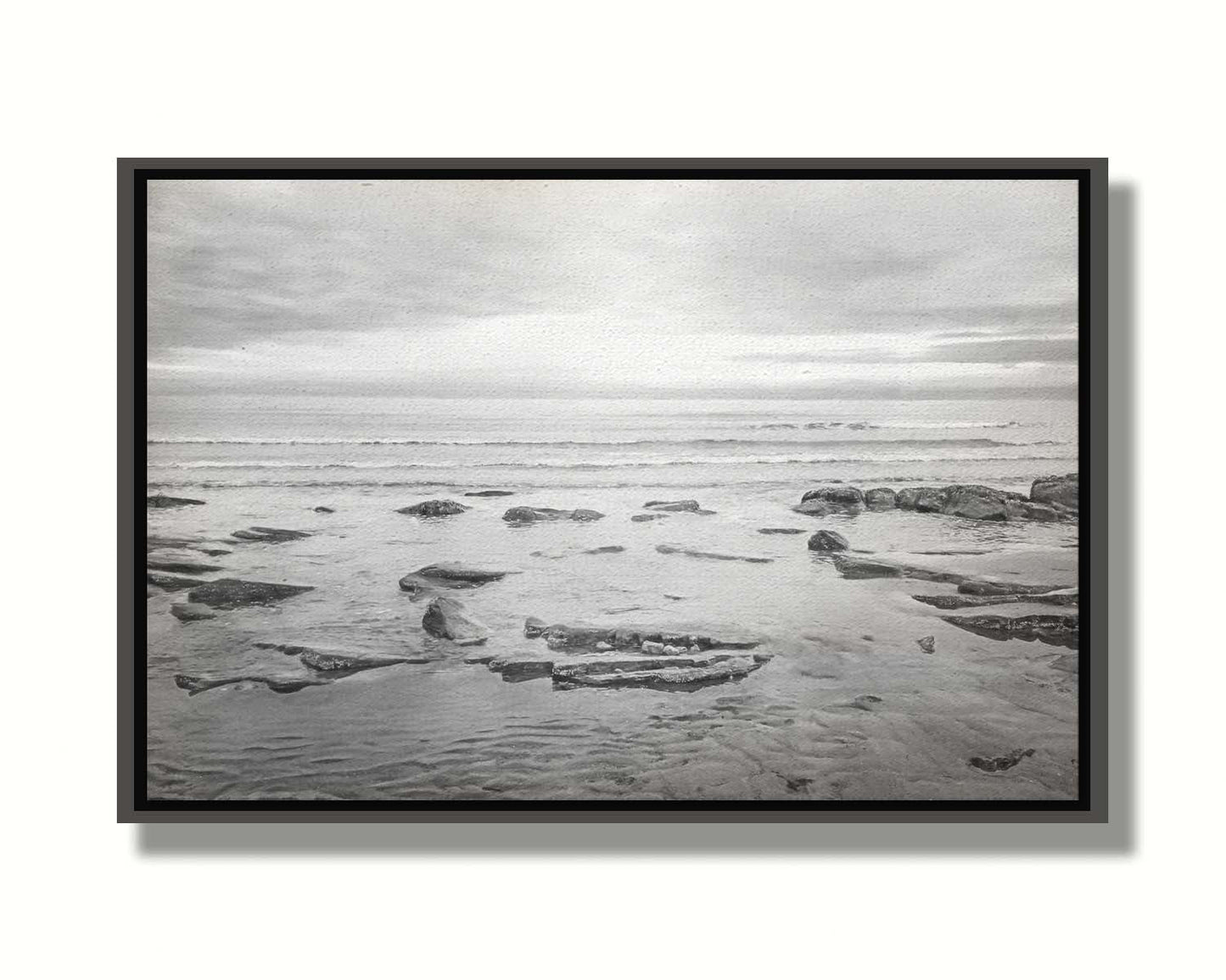 A grayscale photograph of the rocky shore of the North Sea at low tide. The sun peeks over the horizon through the partly cloudy sky. Printed on canvas in a float frame.