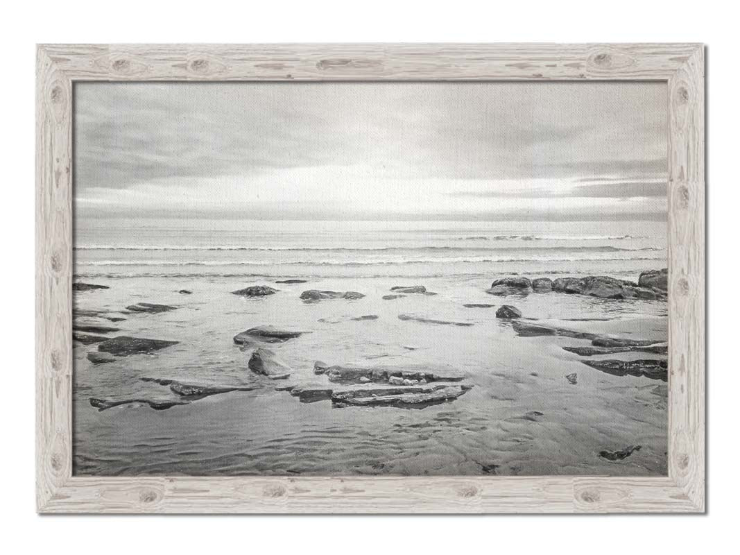 A grayscale photograph of the rocky shore of the North Sea at low tide. The sun peeks over the horizon through the partly cloudy sky. Printed on canvas and framed.