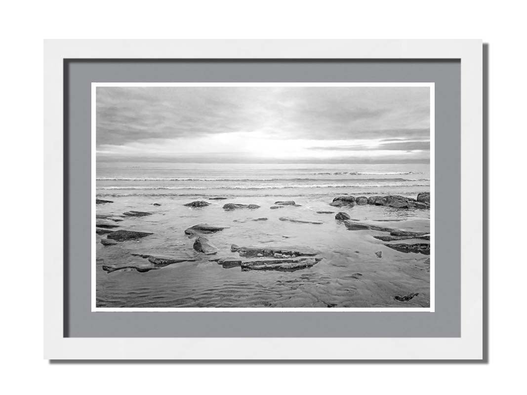 A grayscale photograph of the rocky shore of the North Sea at low tide. The sun peeks over the horizon through the partly cloudy sky. Printed on paper, matted, and framed.