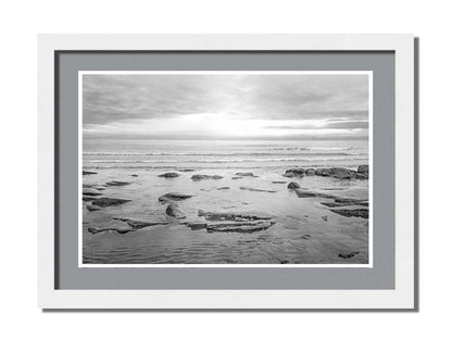 A grayscale photograph of the rocky shore of the North Sea at low tide. The sun peeks over the horizon through the partly cloudy sky. Printed on paper, matted, and framed.