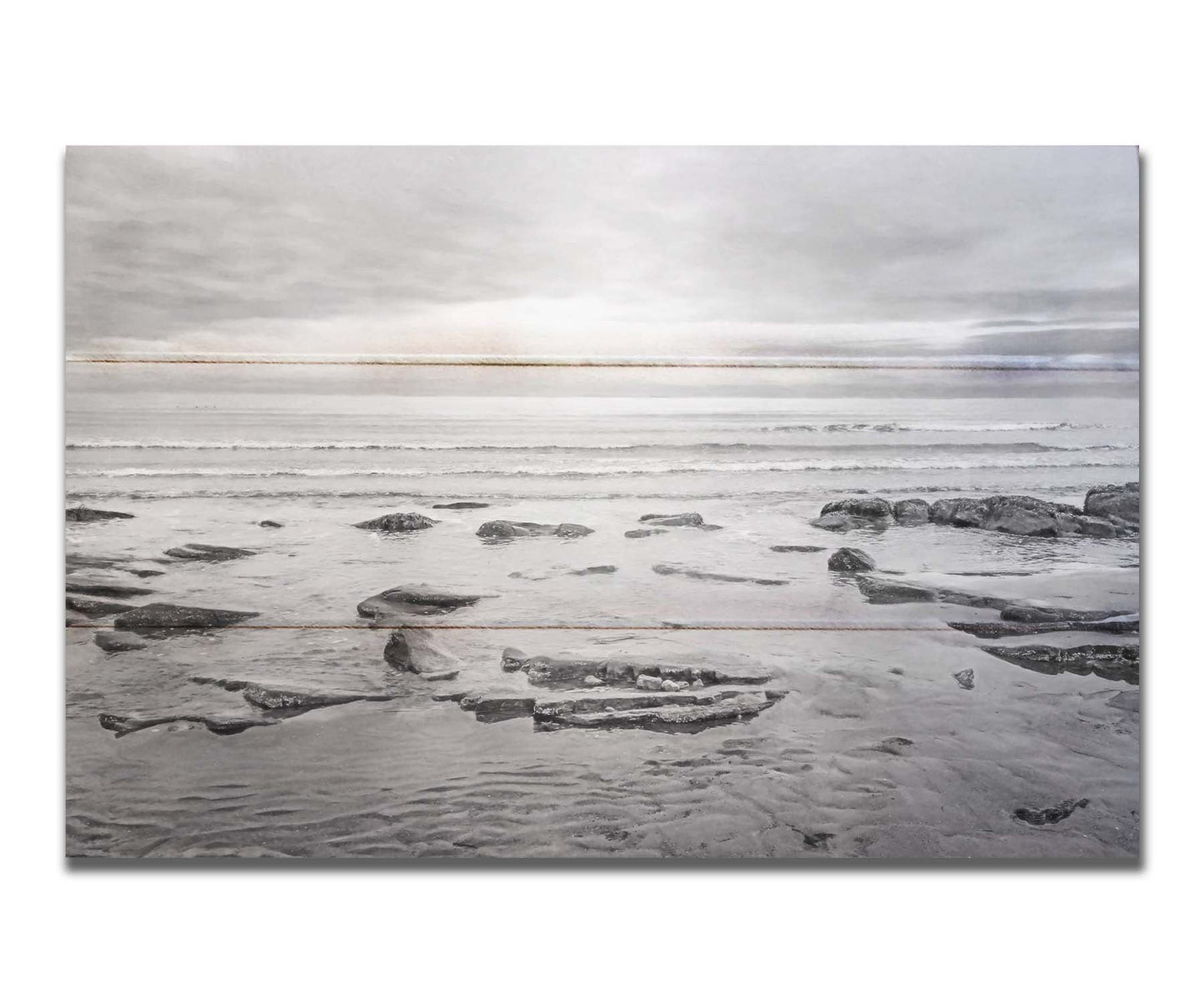 A grayscale photograph of the rocky shore of the North Sea at low tide. The sun peeks over the horizon through the partly cloudy sky. Printed on a box board.