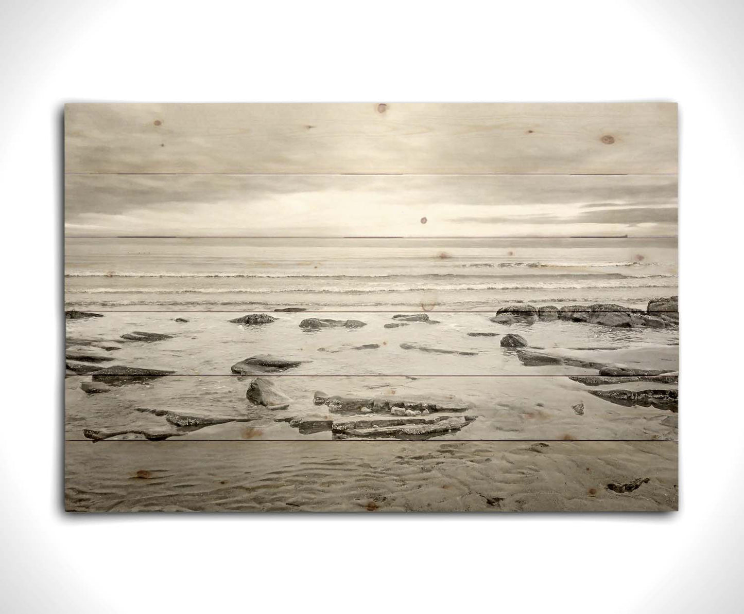 A grayscale photograph of the rocky shore of the North Sea at low tide. The sun peeks over the horizon through the partly cloudy sky. Printed on a wood pallet.