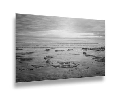 A grayscale photograph of the rocky shore of the North Sea at low tide. The sun peeks over the horizon through the partly cloudy sky. Printed on metal.