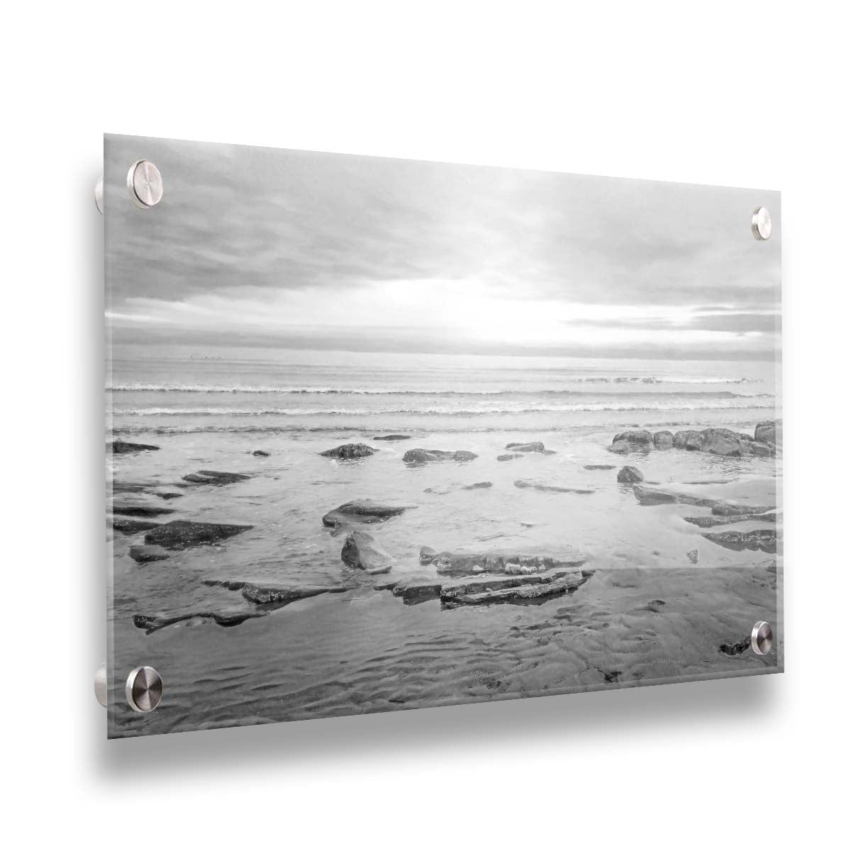 A grayscale photograph of the rocky shore of the North Sea at low tide. The sun peeks over the horizon through the partly cloudy sky. Printed on acrylic.