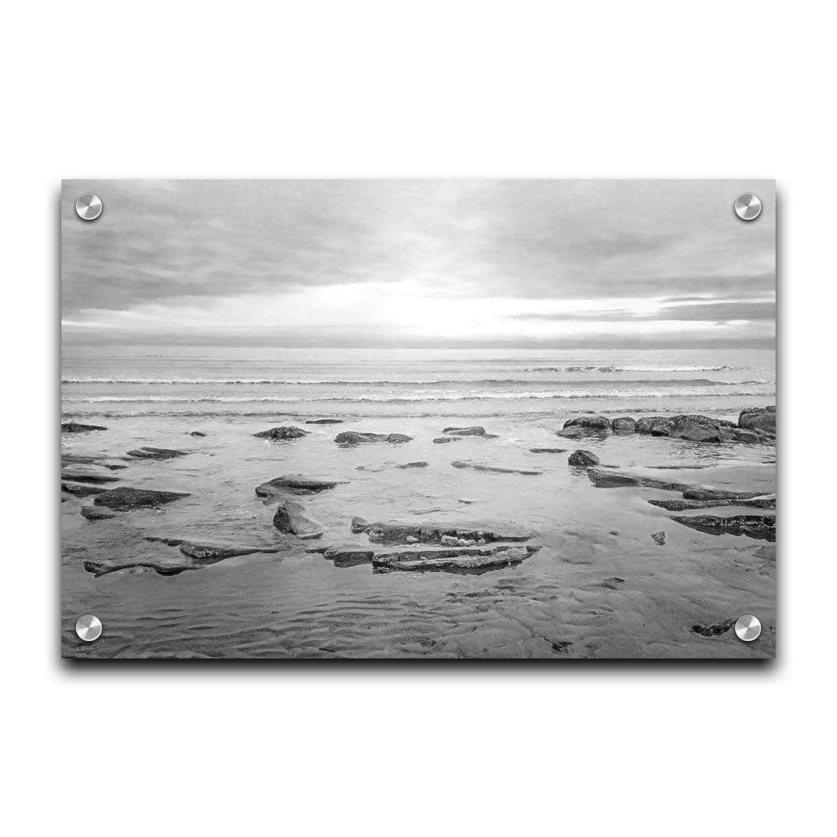 A grayscale photograph of the rocky shore of the North Sea at low tide. The sun peeks over the horizon through the partly cloudy sky. Printed on acrylic.