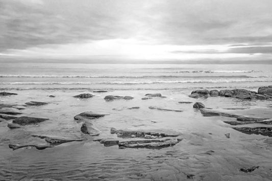 A grayscale photograph of the rocky shore of the North Sea at low tide. The sun peeks over the horizon through the partly cloudy sky.