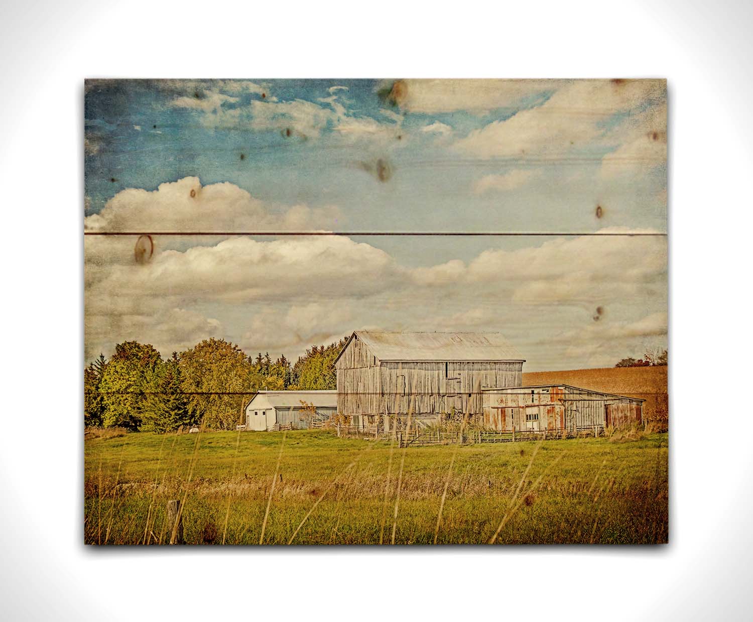 A photo of several barns, their associated pastures, and fields. The early signs of fall are present in the steadily changing leaves and crops. The sky above is wide, blue, and populated with fluffy clouds. Printed on a wood pallet.