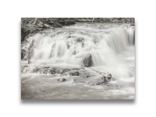 A grayscale photograph of a small river waterfall, only a few feet high. The use of a slow shutter speed technique gives the water a soft, gentle appearance. Printed on canvas.