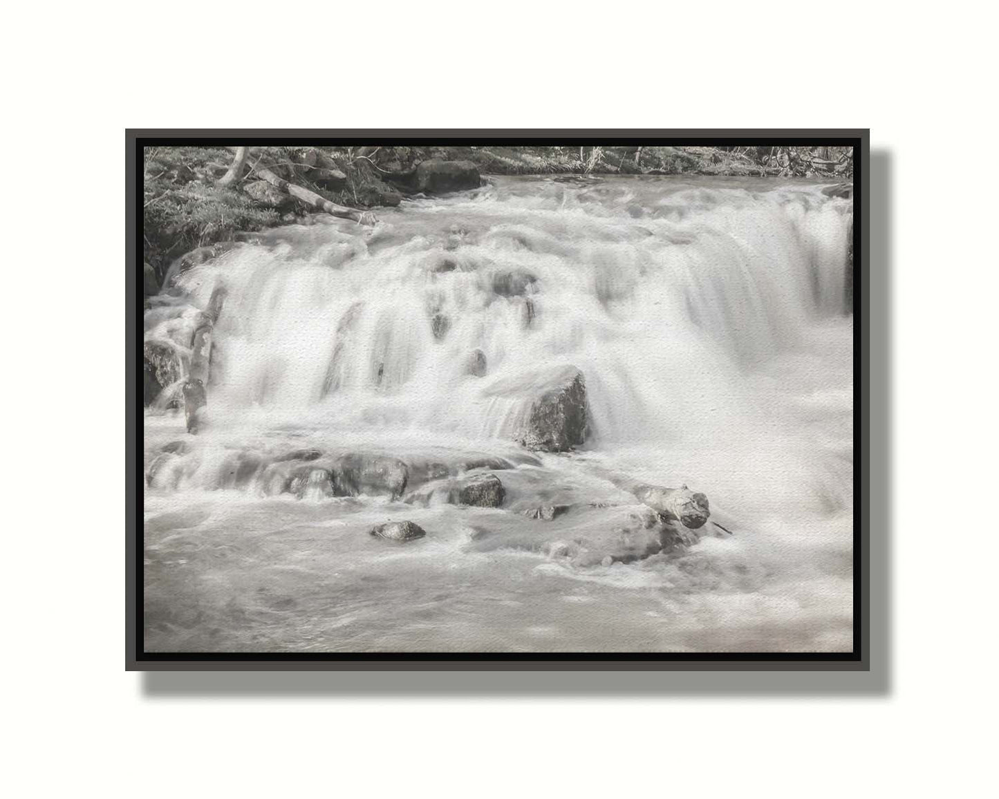 A grayscale photograph of a small river waterfall, only a few feet high. The use of a slow shutter speed technique gives the water a soft, gentle appearance. Printed on canvas in a float frame.