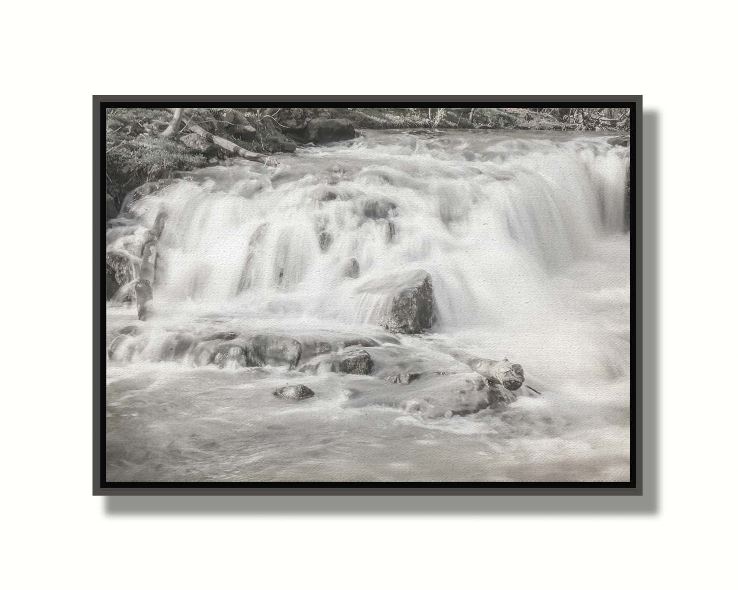 A grayscale photograph of a small river waterfall, only a few feet high. The use of a slow shutter speed technique gives the water a soft, gentle appearance. Printed on canvas in a float frame.