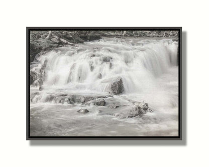 A grayscale photograph of a small river waterfall, only a few feet high. The use of a slow shutter speed technique gives the water a soft, gentle appearance. Printed on canvas in a float frame.