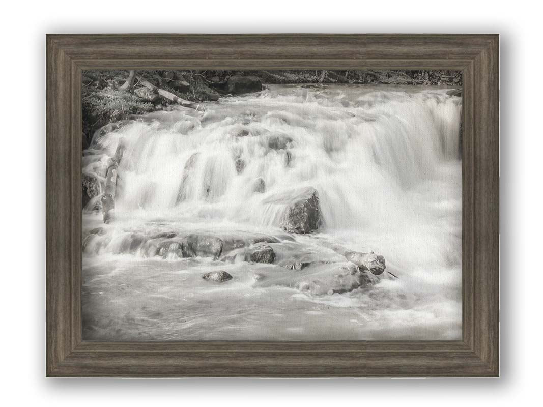 A grayscale photograph of a small river waterfall, only a few feet high. The use of a slow shutter speed technique gives the water a soft, gentle appearance. Printed on canvas and framed.