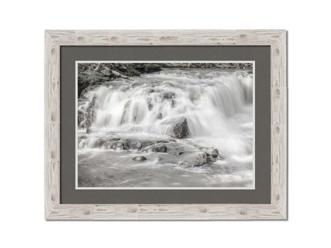 A grayscale photograph of a small river waterfall, only a few feet high. The use of a slow shutter speed technique gives the water a soft, gentle appearance. Printed on paper, matted, and framed.