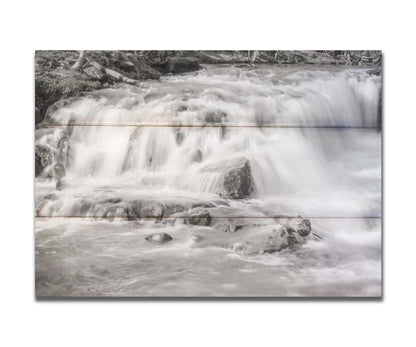 A grayscale photograph of a small river waterfall, only a few feet high. The use of a slow shutter speed technique gives the water a soft, gentle appearance. Printed on a box board.