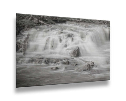 A grayscale photograph of a small river waterfall, only a few feet high. The use of a slow shutter speed technique gives the water a soft, gentle appearance. Printed on metal.