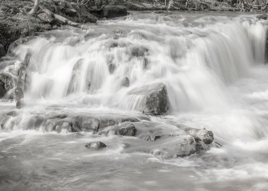 A grayscale photograph of a small river waterfall, only a few feet high. The use of a slow shutter speed technique gives the water a soft, gentle appearance. 
