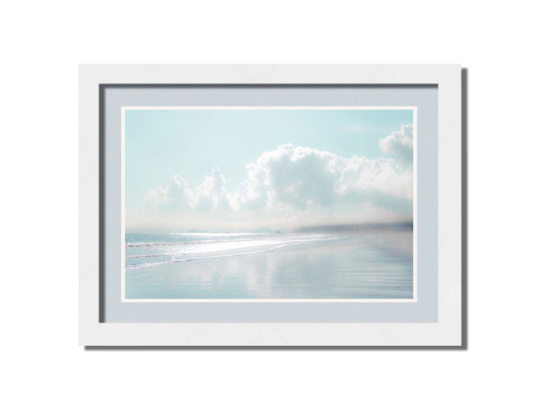 A photo of a beach on a bright but foggy day. The light reflects on the wet sand with the color of the sky,, adding to the dreamlike appearance. A city can be seen through the fog in the distance. Printed on paper, matted, and framed.