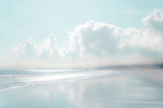 A photo of a beach on a bright but foggy day. The light reflects on the wet sand with the color of the sky, adding to the dreamlike appearance. A city can be seen through the fog in the distance. 