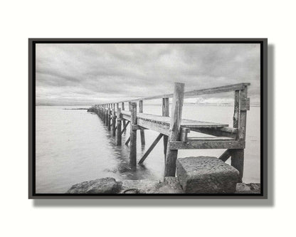 A grayscale photograph of an old wooden pier in Scotland, stretching away from the rocky shore into the waters. Printed on canvas in a float frame.