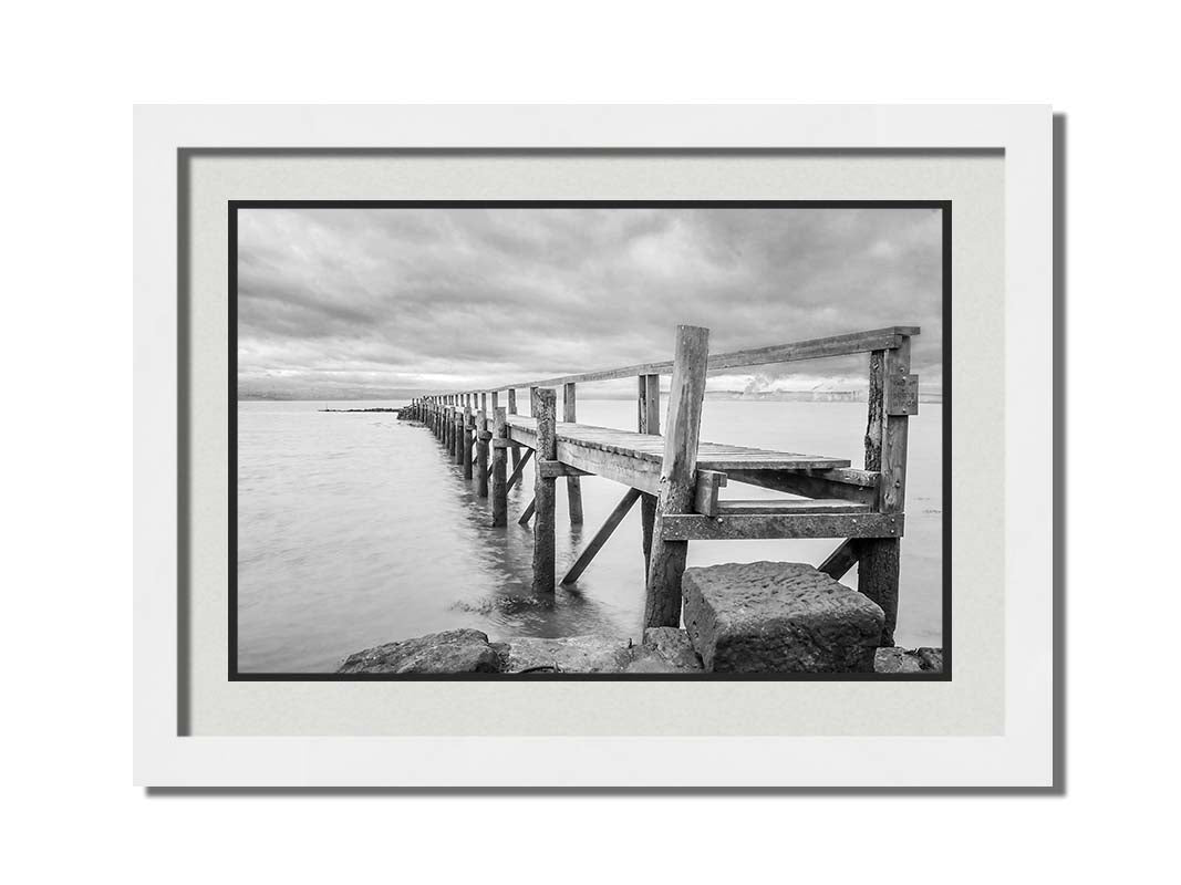 A grayscale photograph of an old wooden pier in Scotland, stretching away from the rocky shore into the waters. Printed on paper, matted, and framed.