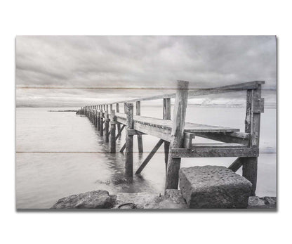 A grayscale photograph of an old wooden pier in Scotland, stretching away from the rocky shore into the waters. Printed on a box board.