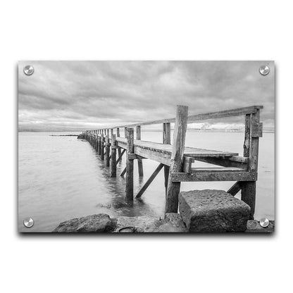 A grayscale photograph of an old wooden pier in Scotland, stretching away from the rocky shore into the waters. Printed on acrylic.