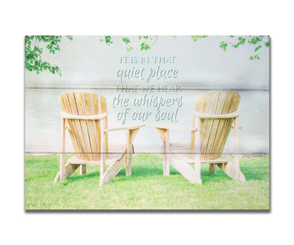 A photo of two wood lawn chairs, sitting on bright green grass, facing a lake. It is overlaid with the phrase, "It is in that quiet place that we hear the whispers of our soul." Printed on a box board.