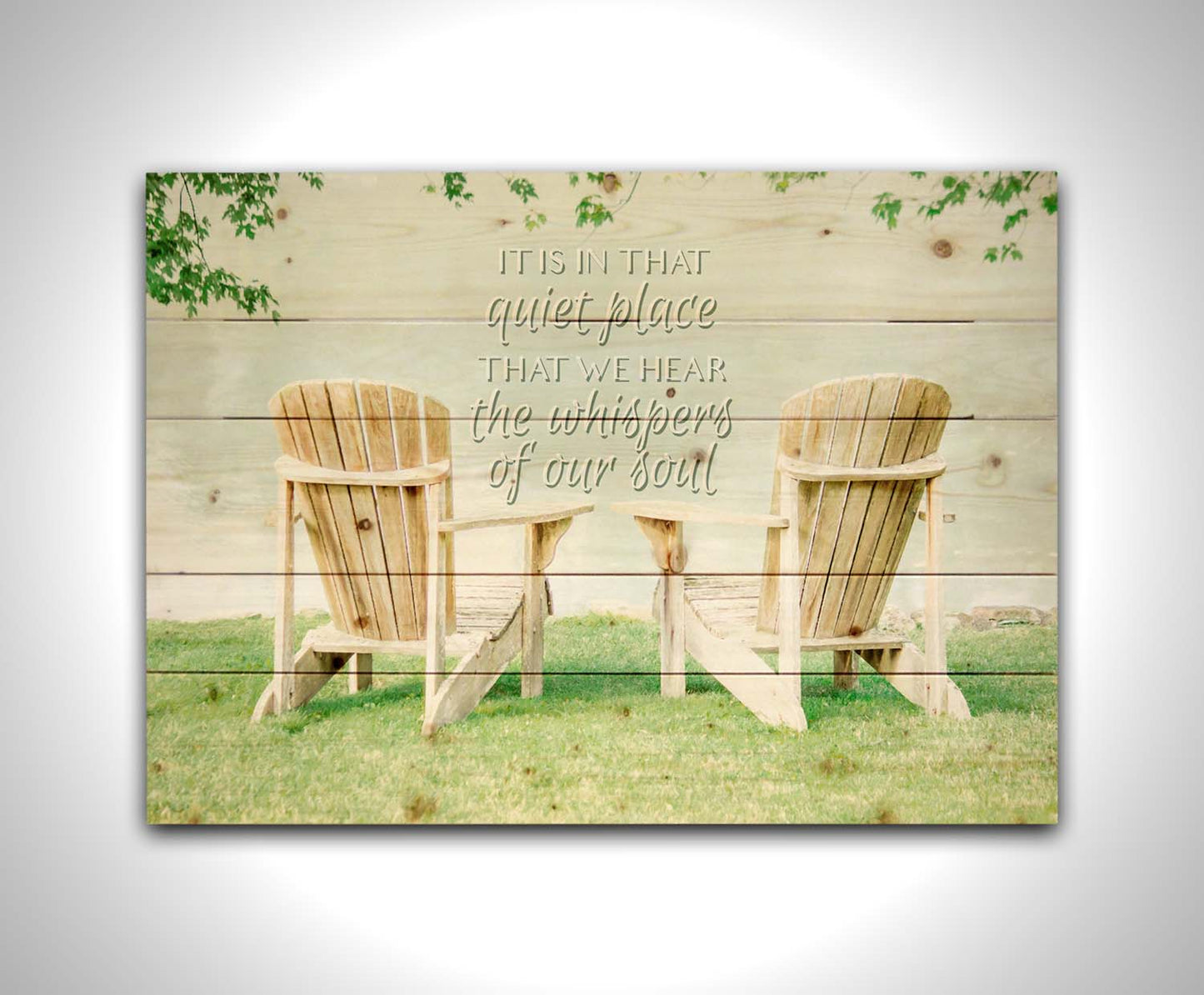 A photo of two wood lawn chairs, sitting on bright green grass, facing a lake. It is overlaid with the phrase, "It is in that quiet place that we hear the whispers of our soul." Printed on a wood pallet.