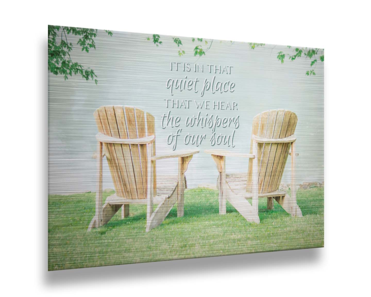 A photo of two wood lawn chairs, sitting on bright green grass, facing a lake. It is overlaid with the phrase, "It is in that quiet place that we hear the whispers of our soul." Printed on metal.