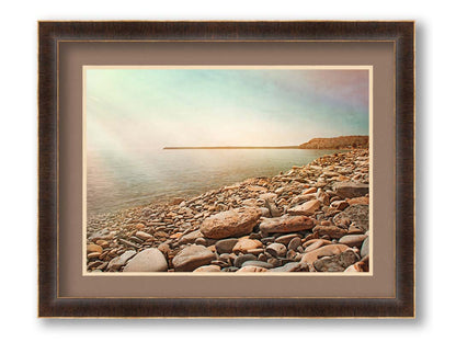 A warm-colored photograph of a rocky beach in the sunlight, bathed in a spectrum of colors. Printed on paper, matted, and framed.