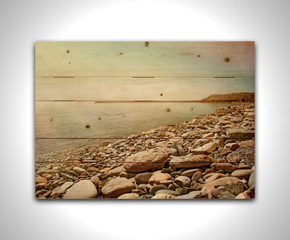 A warm-colored photograph of a rocky beach in the sunlight, bathed in a spectrum of colors. Printed on a wood pallet.