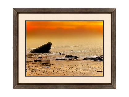 A photo on a beach, looking out at rocks jutting out from the sea against an orange sunset. Printed on paper, matted, and framed.