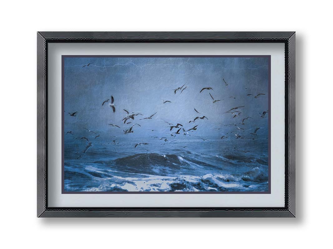 An edited photo of a flock of seagulls swarming over the sea in search of food, as the sky darkens and the waves pick up with an oncoming storm. Printed on paper, matted, and framed.