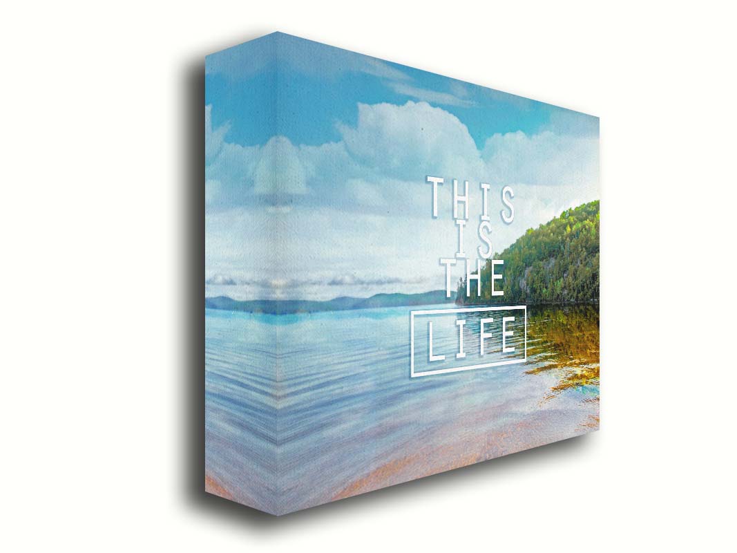 A photograph taken from the shore, looking out toward a forested coast. The water reflects the blue sky and gentle clouds. The phrase "This is the life" is overlaid across the center of the photo in white. Printed on canvas.