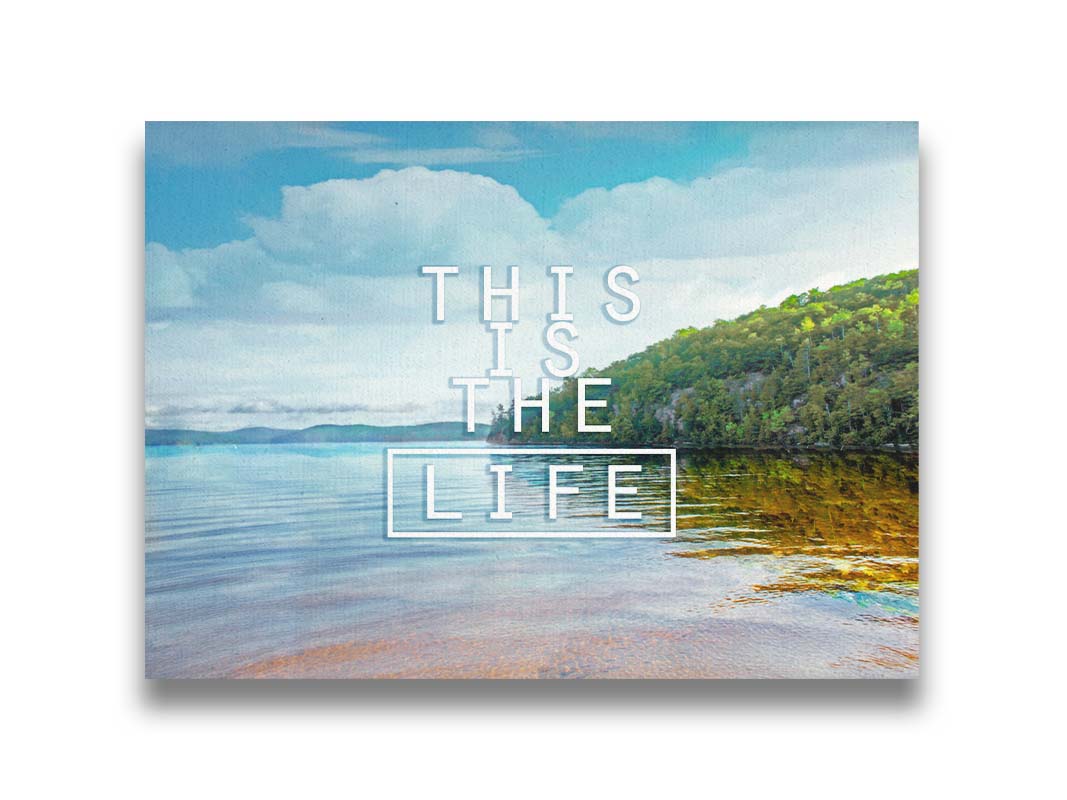 A photograph taken from the shore, looking out toward a forested coast. The water reflects the blue sky and gentle clouds. The phrase "This is the life" is overlaid across the center of the photo in white. Printed on canvas.