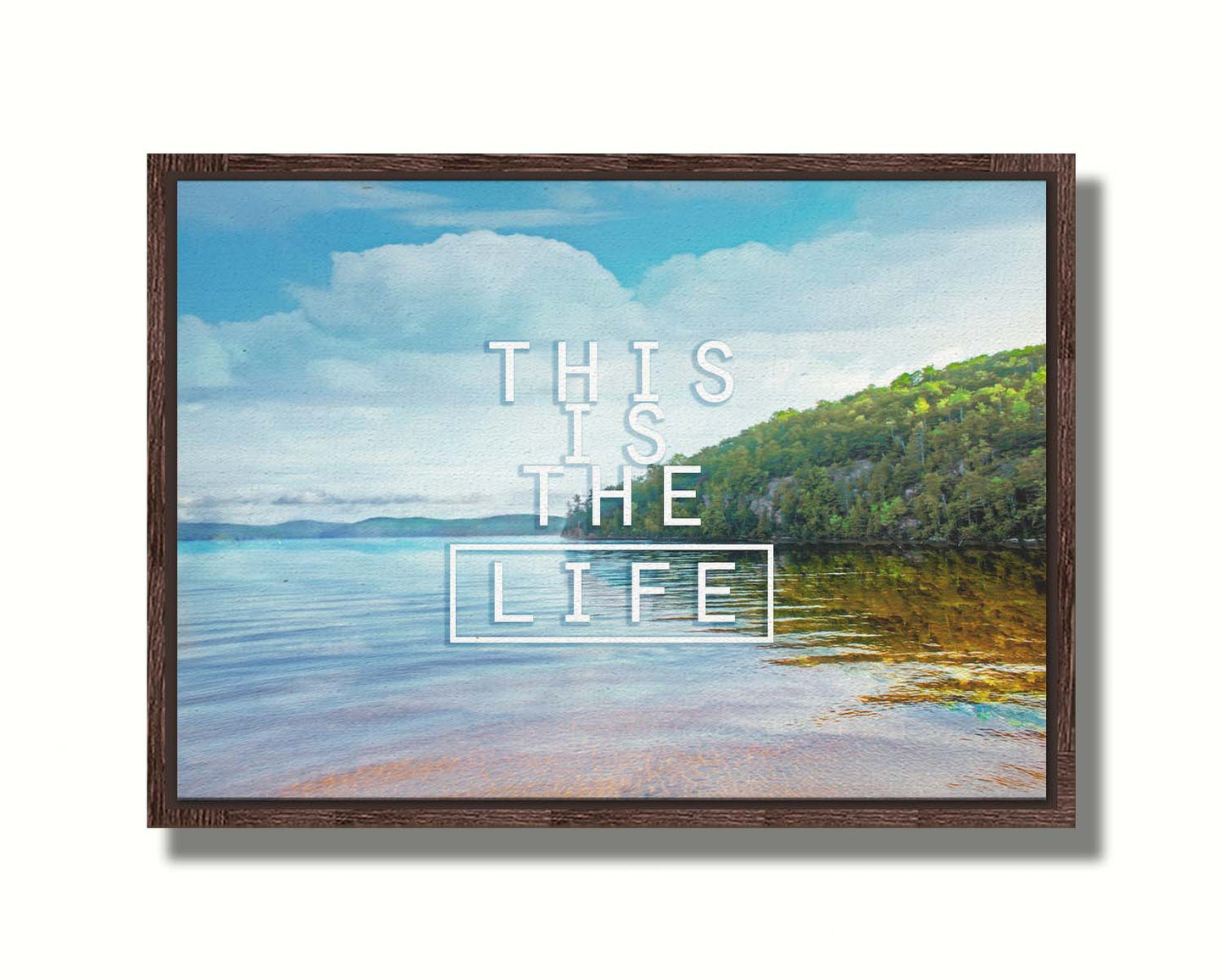 A photograph taken from the shore, looking out toward a forested coast. The water reflects the blue sky and gentle clouds. The phrase "This is the life" is overlaid across the center of the photo in white. Printed on canvas in a float frame.