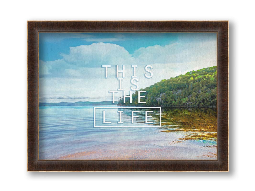 A photograph taken from the shore, looking out toward a forested coast. The water reflects the blue sky and gentle clouds. The phrase "This is the life" is overlaid across the center of the photo in white. Printed on canvas and framed.