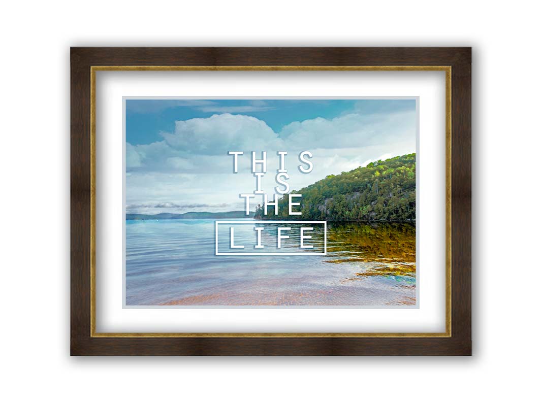 A photograph taken from the shore, looking out toward a forested coast. The water reflects the blue sky and gentle clouds. The phrase "This is the life" is overlaid across the center of the photo in white. Printed on paper, matted, and framed.