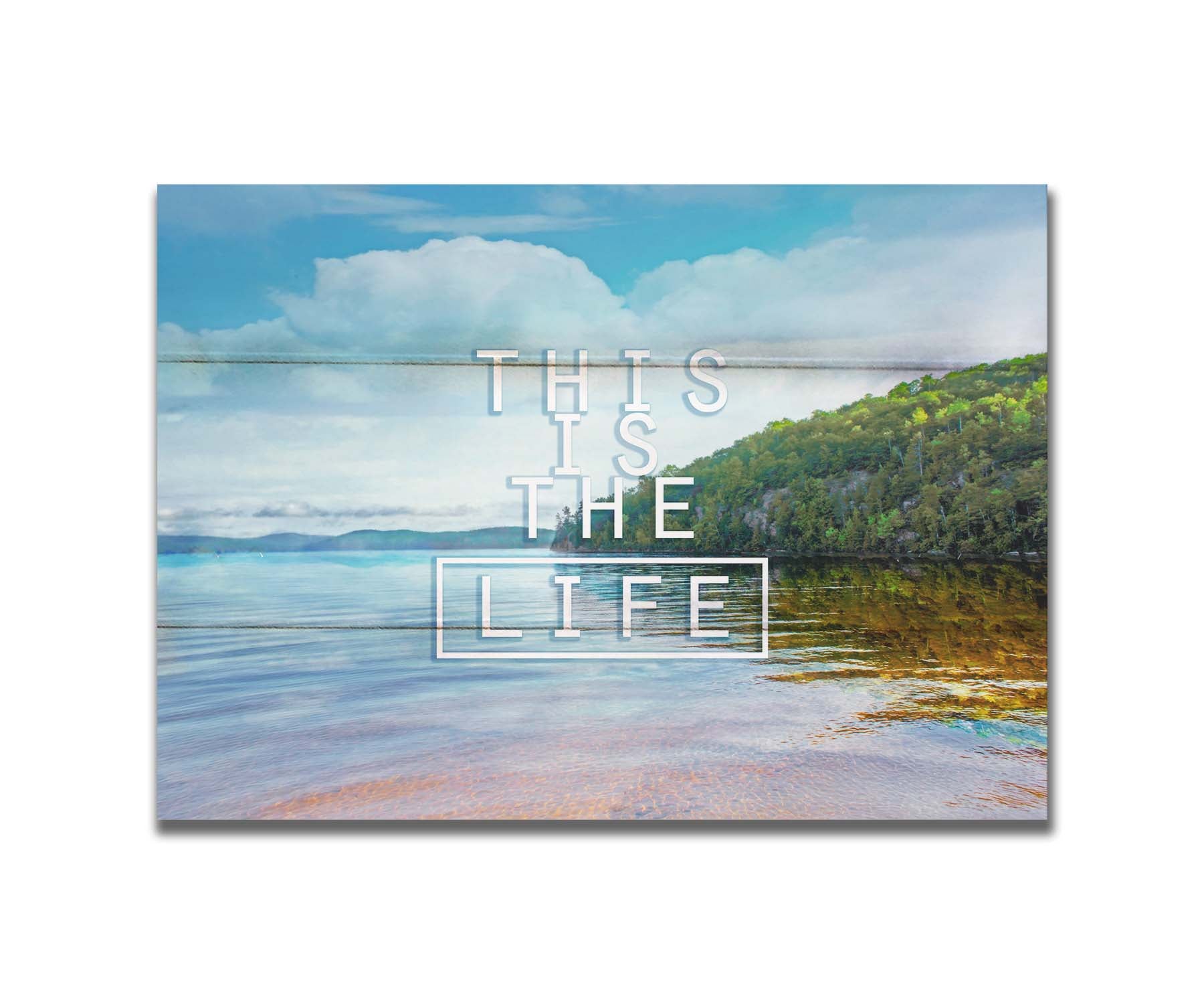 A photograph taken from the shore, looking out toward a forested coast. The water reflects the blue sky and gentle clouds. The phrase "This is the life" is overlaid across the center of the photo in white. Printed on a box board.