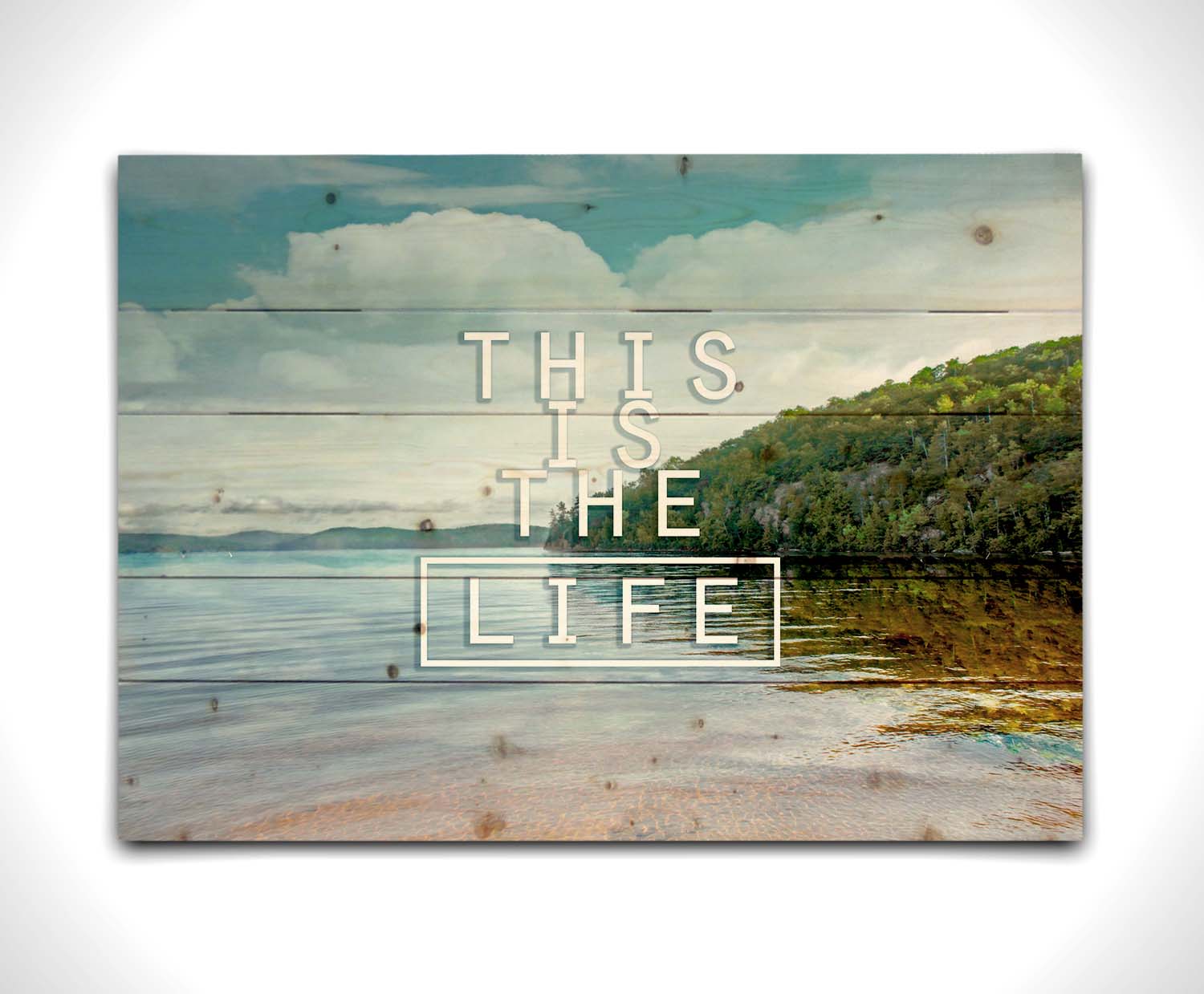 A photograph taken from the shore, looking out toward a forested coast. The water reflects the blue sky and gentle clouds. The phrase "This is the life" is overlaid across the center of the photo in white. Printed on a wood pallet.