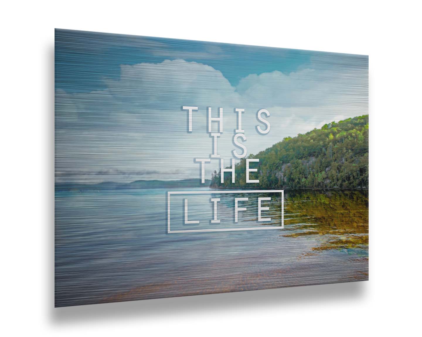 A photograph taken from the shore, looking out toward a forested coast. The water reflects the blue sky and gentle clouds. The phrase "This is the life" is overlaid across the center of the photo in white. Printed on metal.