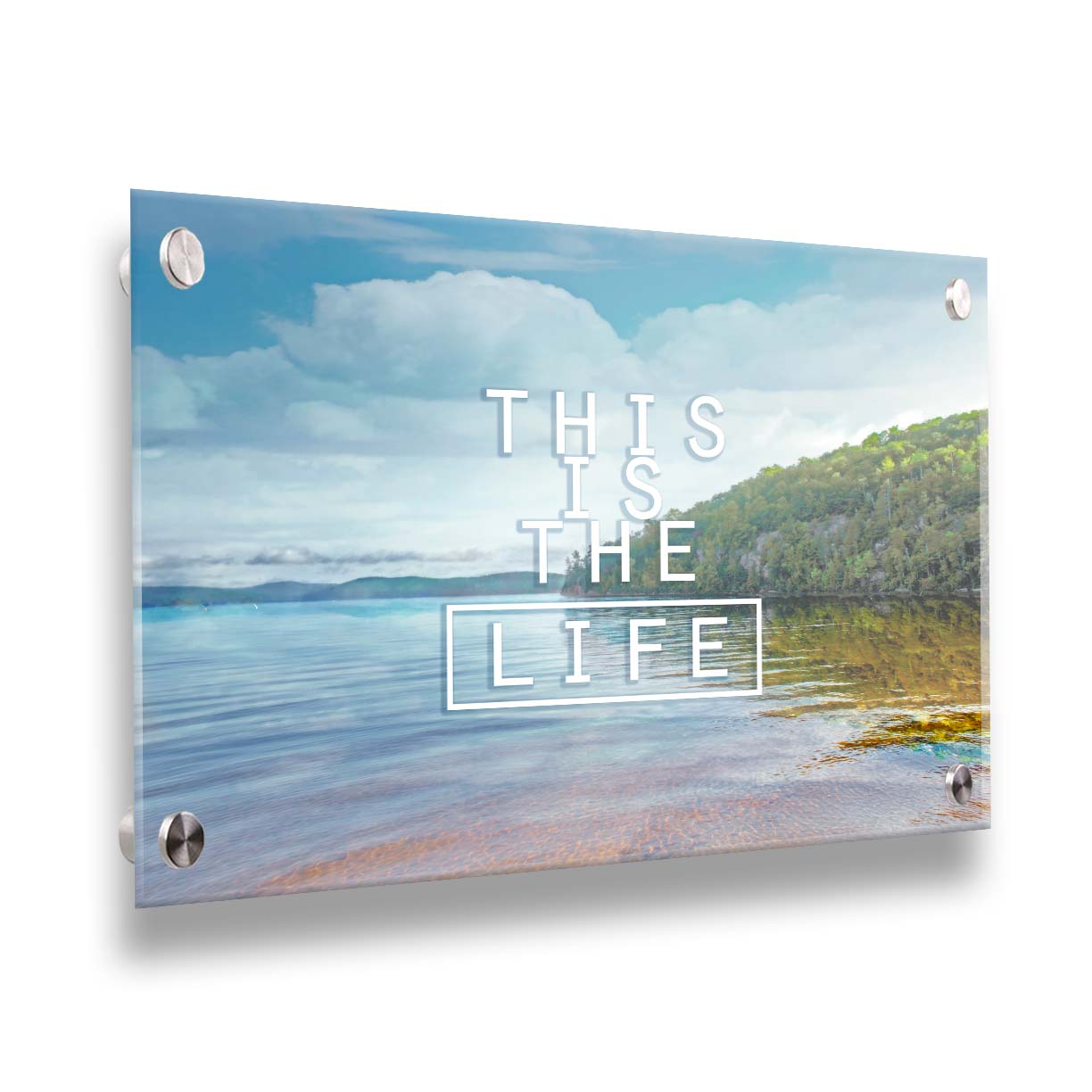 A photograph taken from the shore, looking out toward a forested coast. The water reflects the blue sky and gentle clouds. The phrase "This is the life" is overlaid across the center of the photo in white. Printed on acrylic.