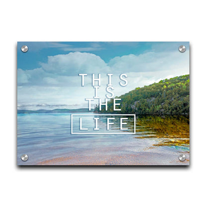 A photograph taken from the shore, looking out toward a forested coast. The water reflects the blue sky and gentle clouds. The phrase "This is the life" is overlaid across the center of the photo in white. Printed on acrylic.