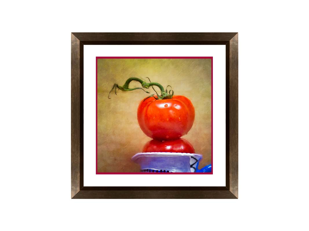 A photograph of bright red tomatoes stacked inside a blue and white ceramic cup, set against a textured neutral background. Printed on paper, matted, and framed.