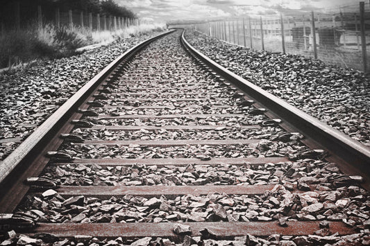 A photo looking down train tracks in black and white, with tinges of red-brown spot color.