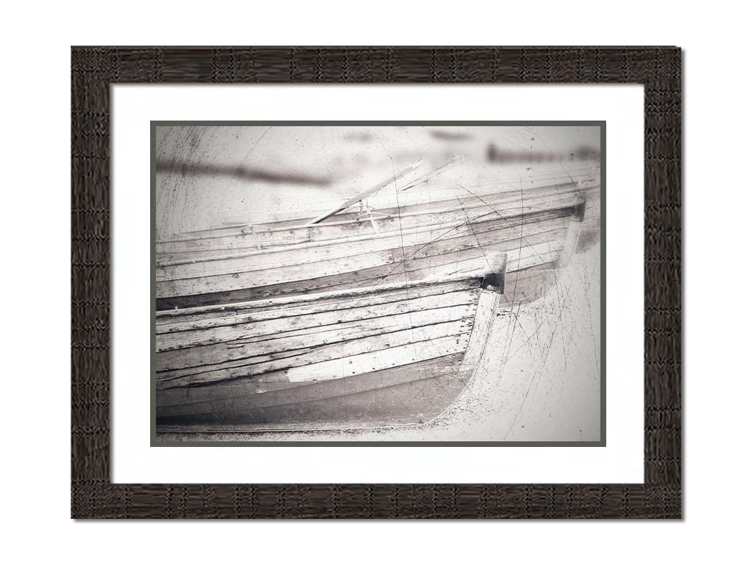 A grayscale photo of two rowboats on the sand. It is overlayed with a scratchy weathered texture. Printed on paper, matted, and framed.