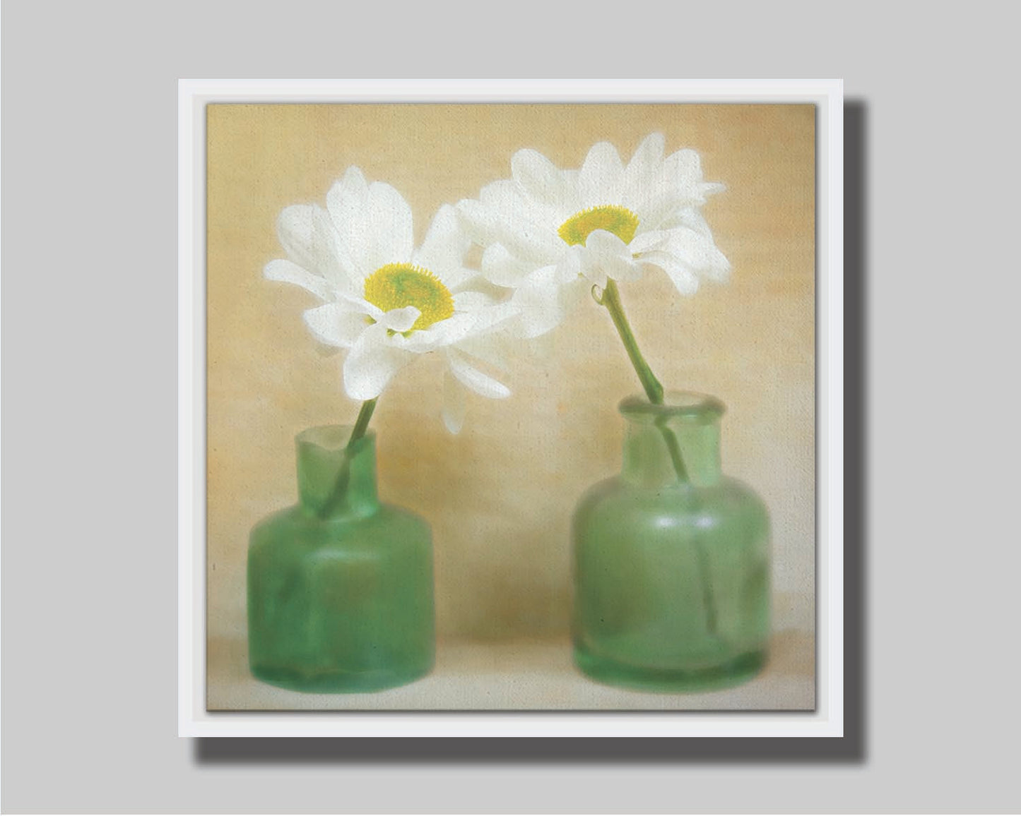 A photo of two white flowers in green glass jars, sitting against one another against a tan backdrop. Printed on canvas in a float frame.