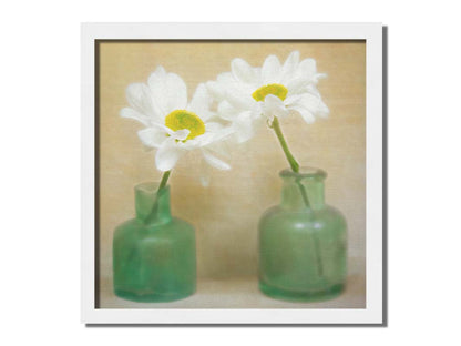 A photo of two white flowers in green glass jars, sitting against one another against a tan backdrop. Printed on canvas and framed.