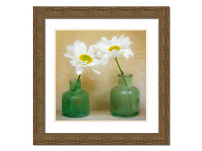 A photo of two white flowers in green glass jars, sitting against one another against a tan backdrop. Printed on paper, matted, and framed.